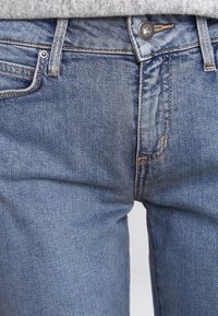 Los jeans de mezclilla azul claro presentan un ajuste slim, costuras visibles, un diseño de cinco bolsillos y un cierre de botón metálico en la cintura.