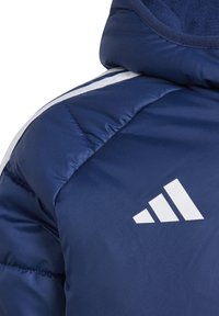 Navy blue padded jacket with white stripe details and an embroidered white logo on the shoulder. Smooth, shiny fabric texture.