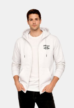 Ung mand iført en hvid hættetrøje med lynlås med teksten "Off Road Club", kombineret med en hvid T-shirt og sorte bukser, hænder i lommerne.