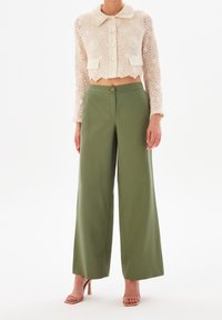 Giacca in crochet di colore crema, tagliata, con colletto rotondo e quattro bottoni, abbinata a pantaloni a vita alta e gamba larga di colore verde oliva.