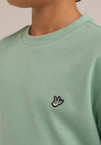 Miętowy sweatshirt wykonany z miękkiego materiału, z okrągłym dekoltem i małym, wyszywanym logo ptaka w czerni i bieli po lewej stronie.
