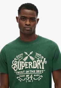 Grøn bomulds T-shirt med et hvidt grafisk design med pile og teksten "SUPERDRY" og "TRUST IN THE BEST", klassisk crew neck stil.