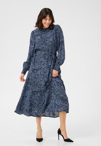 Robe bleu marine à manches longues avec un motif texturé blanc, une taille ceinturée et une jupe fluide, associée à des escarpins noirs.