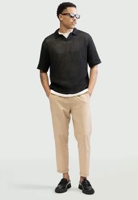 Muž v černém síťovaném svetru s krátkým rukávem, béžových kalhotách, černých loaferkách, slunečních brýlích, s rukama v kapsách, stoj�ící na neutrálním pozadí.