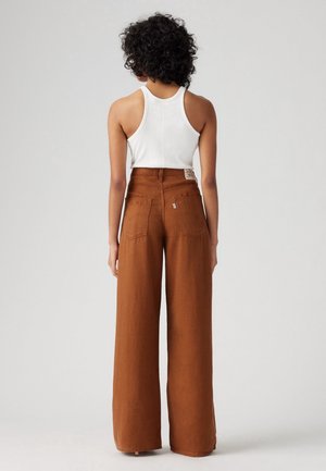 Personne aux cheveux bouclés portant un haut blanc sans manches et un pantalon marron large taille haute, debout de dos sur un fond uni.