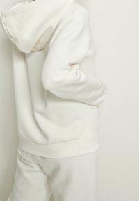 Persoon in een off-white capuchontrui en bijpassende broek met de handen in de zakken van de trui, gezien van achteren.