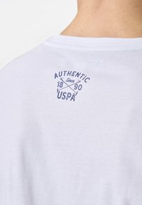 Baltas medvilnės T-shirt su apvaliu kaklu, ant nugaros su jūrinės mėlynos spalvos spaudiniu, kuriame parašyta "AUTENTIŠKAS Nuo 1890 m. USPA".
