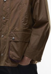 Brown waxed jacket with metal snap buttons and a side pocket. The fabric has a smooth texture, featuring purple accents near the cuffs.