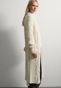 Larga chaqueta de punto en color crema con patrones de trenzas, de apertura frontal y puños acanalados. Presenta una cintura atada y aberturas laterales para facilitar el movimiento.