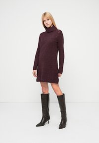 Burgunderfarget strikket kjole med turtleneck og kabelmønster, med lange ermer og sidespalte. Kombinert med knelange svarte skinn-støvler.