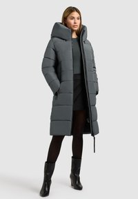 Grå pufferjakke med en høj, struktureret hætte, der har quiltede rektangler og skjulte lommer. Båret over en mørk bluse og nederdel.