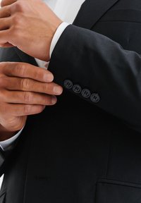 Czarna dopasowana marynarka o gładkiej fakturze z trzema czarnymi guzikami przy mankietach, z białą koszulą widoczną poniżej.