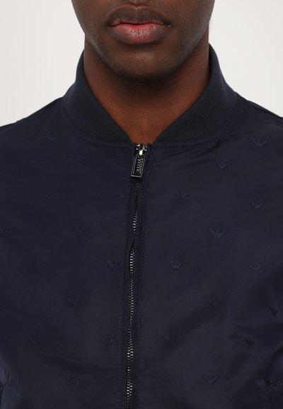 Navy bomber jacket with a zip front, ribbed collar, and embossed logo pattern throughout the fabric. Smooth texture and lightweight material.