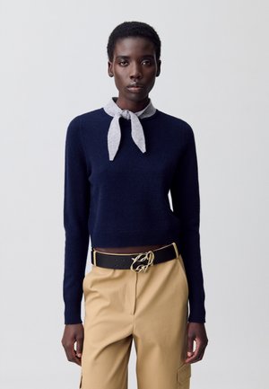 Pull navy à manches longues et cravate gris clair. Porté avec un pantalon beige avec poches et une ceinture noire avec une boucle en matériel doré.