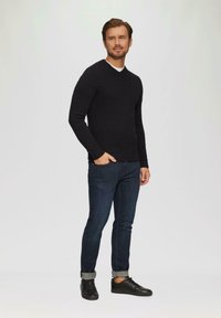 Schwarzer langärmliger Pullover mit V-Ausschnitt, kombiniert mit dunkelblauen Jeans und schwarzen Sneakern, die gekrempelte Bündchen als Detail aufweisen.