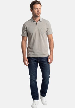 Mand stående iført lysegrå kortærmet polo, mørkeblå jeans og hvide sneakers, kiggende til siden.