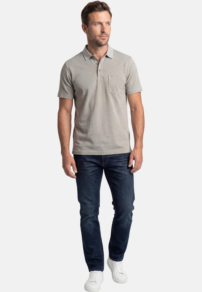 Homme debout portant un polo à manches courtes gris clair, un jean bleu foncé et des baskets blanches, regardant sur le côté.