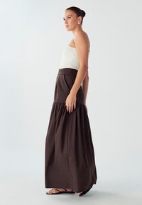 Hnedá maxi sukňa s poschodovým dizajnom, so sťaženým pásom a voľným lemom. Skombinovaná s bielym rebrovaným topom bez ramienok a plochými sandálmi.