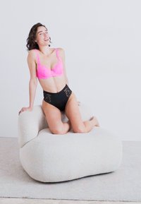 Kurviger, plüschiger Stuhl aus weichem, weißen Stoff; glatte, abgerundete Form. Modell sitzt in einem leuchtend rosa Spitzen-BH und schwarzen hochtaillierten Panties.