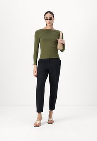 Haut ajusté à manches longues vert olive associé à un pantalon noir ajusté et des sandales beiges. Le mannequin tient un petit sac à main beige.