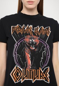 Μαύρο κοντομάνικο μπλούζα με γραφικό σχέδιο ενός ροχαλίσματος τίγρη, με πορτοκαλί λεπτομέρειες, μωβ γραμμές ηλεκτρικής εκκένωσης και έντονο κείμενο "VERSACE JEANS COUTURE".