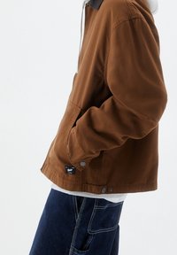 Brązowa kurtka wykonana z miękkiego materiału z czarnym kołnierzem. Posiada przednie kieszenie i zapięcie na napy, noszona na jasnym swetrze.