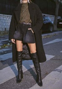 Osoba v černém dlouhém kabátu, olivovém svetr s copánkovým vzorem, černých šortkách s vysokým pasem a černých kozačkách stojí na ulici.