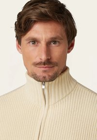 Cremiger, gerippter Pullover mit hohem Kragen und vollem Reißverschluss. Die Textur ist weich mit vertikalen Linien und einem subtilen Glanz auf dem Reißverschluss.