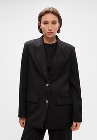 Blazer negru cu un design ajustat, model discret cu puncte, butoane accente argintii, rever cu decupaj și două buzunare frontale. Textură netedă.