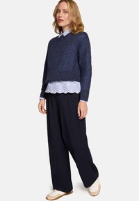 Pull marin en tricot superposé sur un chemisier bleu clair à pois avec ourlet festonné, associé à un pantalon noir à jambes larges et des baskets crème.