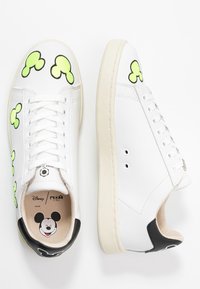 Białe skórzane sneakersy z zielonymi sylwetkami Myszki Mickey, sznurowane, z czarnymi akcentami na pięcie i nadrukowanym logo Myszki Mickey w środku.