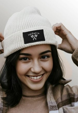 Cream-colored knit beanie with a ribbed texture, featuring a black rectangular patch labeled "valhalla" and two crossed axes.