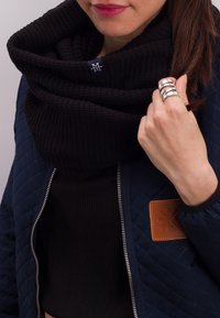 Femme portant une écharpe noire côtelée et une veste matelassée bleu marine avec une bague argentée à la main droite, tenant une mèche de cheveux bruns.