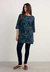 Tunic floreale con maniche a tre quarti di colore blu scuro, caratterizzata da vivaci motivi verdi e arancioni, abbinata a pantaloni skinny scuri e zoccoli marroni.
