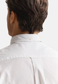 Mann von hinten gesehen mit kurzem, dunklem Haar, der ein weißes Hemd mit Knopfleiste und Kragen vor einfarbigem Hintergrund trägt.