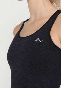 Czarny sportowy top bez rękawów o teksturowanym wykończeniu, z okrągłym dekoltem i małym okrągłym logo w kolorze srebrnym po lewej stronie u góry.