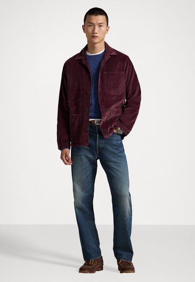 Burgundy corduroy jacket with a single pocket, worn over a blue sweater, paired with blue jeans and brown lace-up shoes.