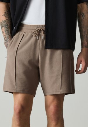 Mann trägt braune, strukturierte Shorts mit Kordelzug und Taschen sowie ein teilweise geöffnetes schwarzes Hemd über einem weißen Top, das tätowierte Arme zeigt.