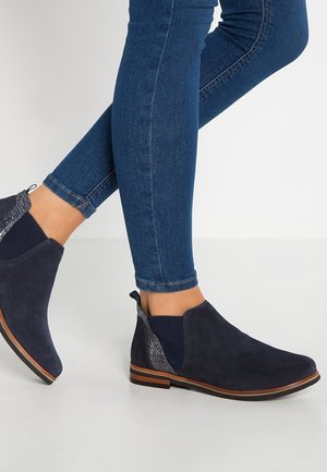 Personne portant des bottines en daim bleu foncé avec des côtés élastiques et un détail de talon texturé peau de serpent, assorties à un jean bleu ajusté.