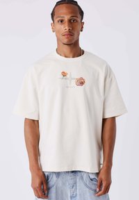 Camiseta de algodón crema con corte oversized, con acentos gráficos florales y un diseño geométrico en el centro. Mangas cortas, escote redondo.