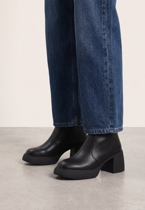 Schwarze Leder-Stiefeletten mit klobigen Absätzen, getragen zu gerade geschnittenen blauen Jeans auf hellem Boden und schlichtem Hintergrund.