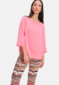 Helles pinkes Langarmoberteil mit einem runden Ausschnitt und Glockenärmeln, kombiniert mit einer multicoloren Zickzack-musternden Hose.