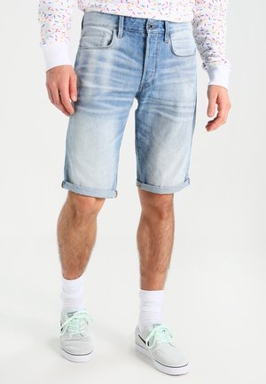 Homme portant un short en jean bleu clair retroussé, des chaussettes blanches, des baskets blanches et une chemise blanche à manches longues avec un motif de confettis colorés.