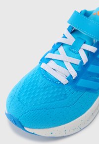 Pantof sport albastru deschis cu șireturi albe, partea superioară din material texturat și talpă albă cu pete. Are o curea peste laba piciorului.