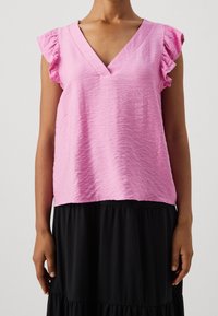 Kvinna som bär en ljusrosa texturerad V-ringad ärmlös blus med volangärmar och en svart lager-på-lager kjol.