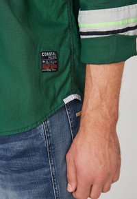 Chemise verte à manches longues avec une coupe carrée, dotée d'une étiquette patch et d'accents rayés en gris, blanc et vert néon sur les manches.