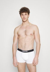 Baltas kokvilnas bokseršorti ar melnu Levi's logotipa jostas daļu, cieši pieguļošs dizains un gluda tekstūra. Modelis ir bez krekla un stāv.