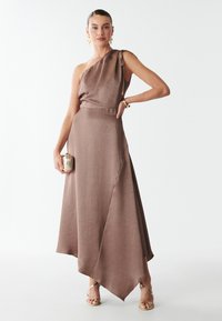 Abito in raso marrone con spallina unica e orlo asimmetrico, dotato di una cintura larga. Gli accessori includono una clutch dorata e sandali con tacco alto e cinturini.