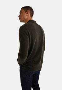 Pull en tricot vert foncé avec un col et des poignets côtelés. Porté avec un jean en denim bleu foncé, affichant une texture lisse. Design sobre.