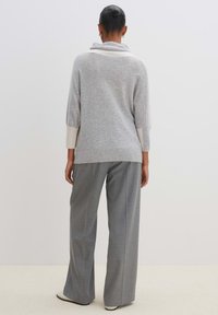 Grau gestrickter Pullover mit Schalkragen, langen Ärmeln und kontrastierenden hellgrauen Akzenten an den Seiten. Kombiniert mit lässigen grauen Hosen.